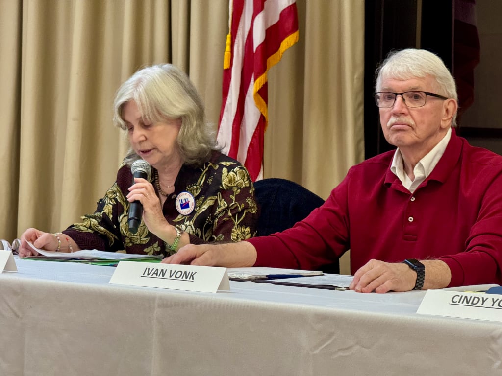 A woman with microphone presenting script at a table next to a white-haired man