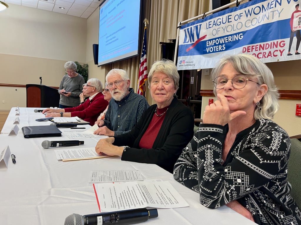 A view of people sitting at a table from the short end with the closest people making interesting faces at the photographer