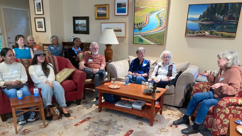 Nine people in a living room setting listening to a woman speaking