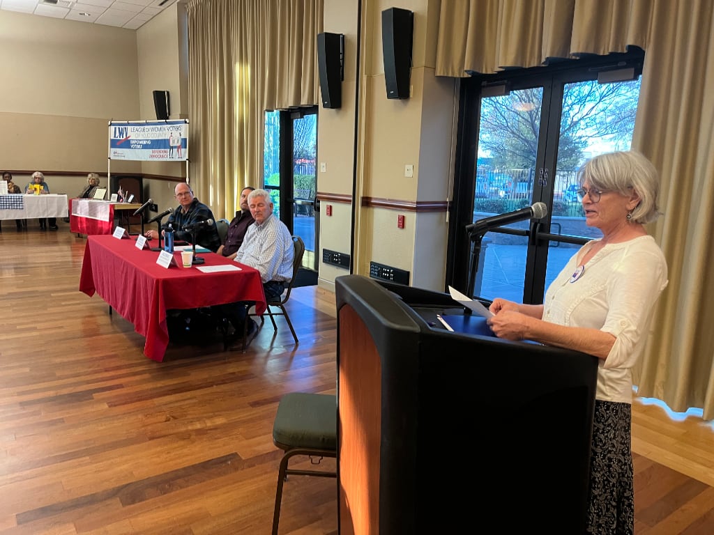 A woman at a podium presenting information about the 3-man panel at a table in the middle distance