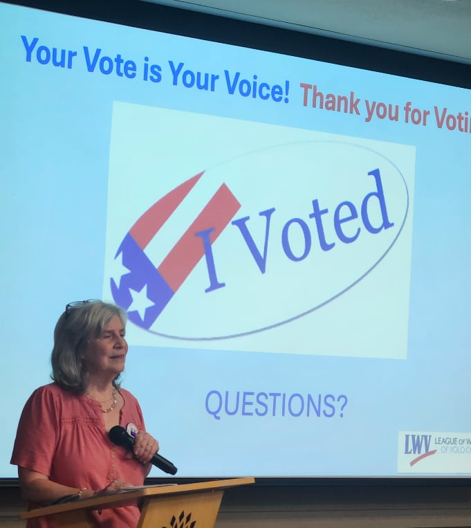 A woman at a podium with a large I Voted sticker on the screen behind her
