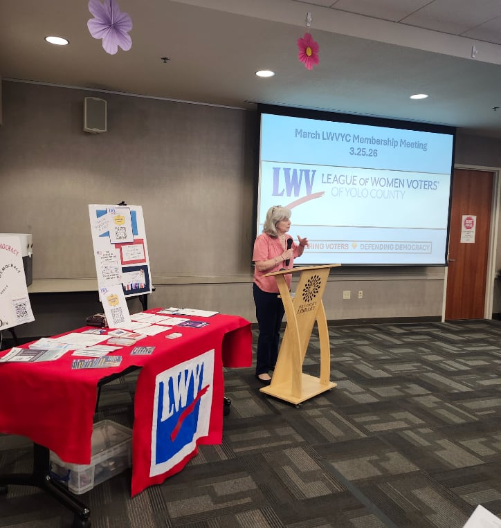 A woman at a podium with a LWV table in the foreground and large presentation screen in the background