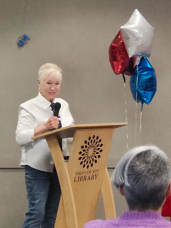 A white-haired woman at a podium with the Yolo County Library logo on the front.