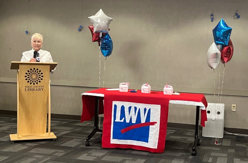 A white-haired woman at a podium next to a table with red-white-blue balloons and LWV banner